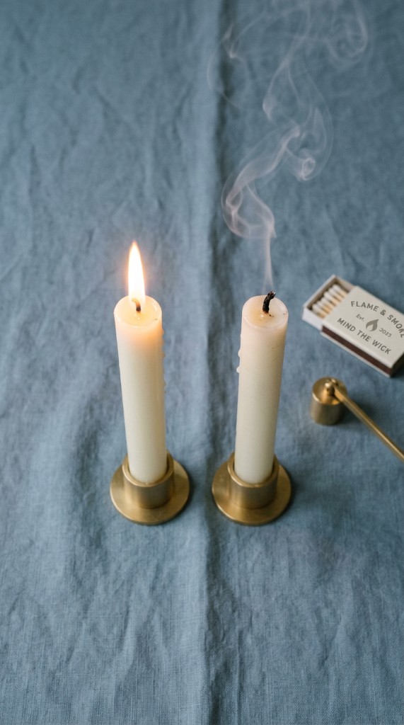 Two white taper candles on blue linen, one lit and one just extinguished with soft smoke rising
